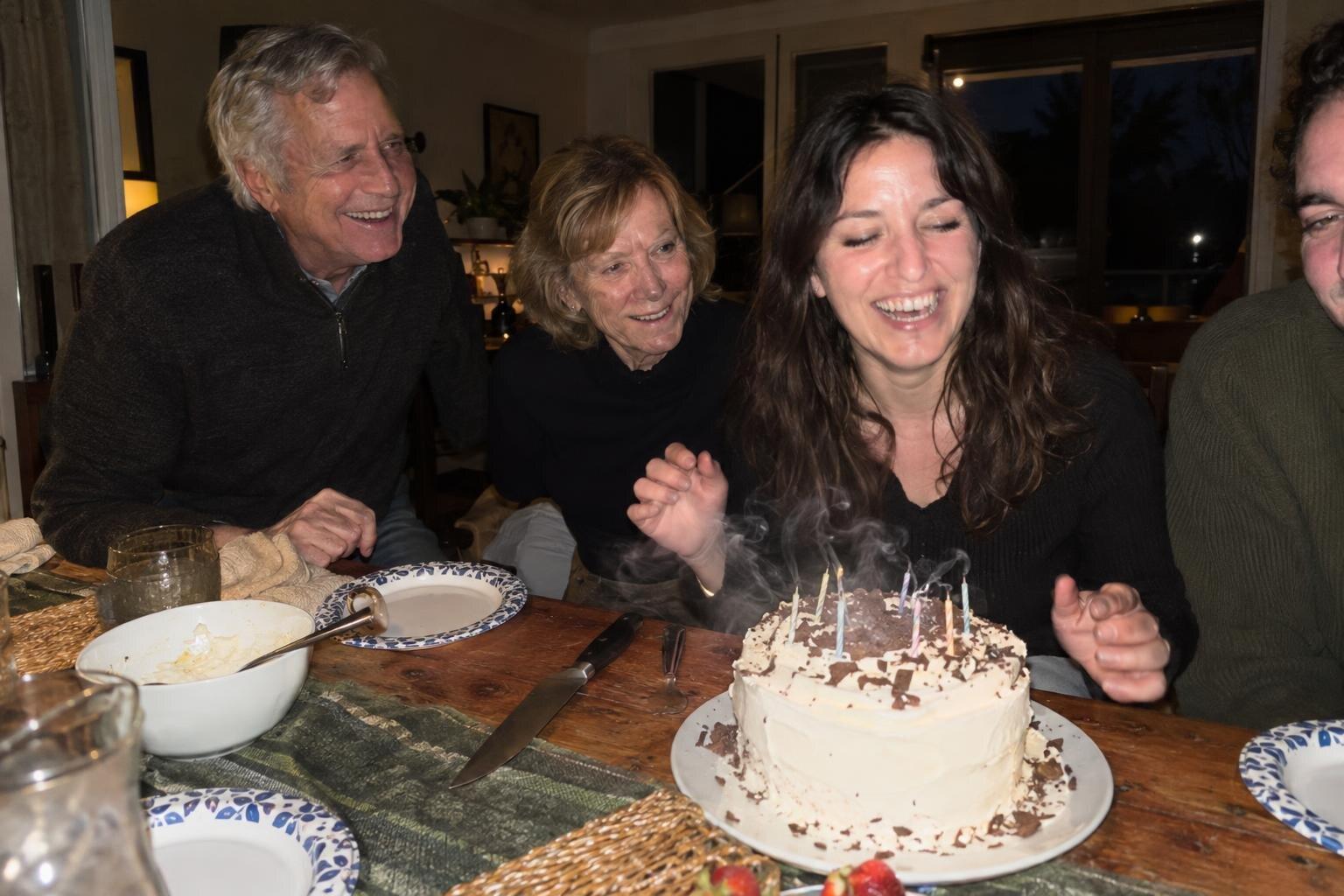 Four adults laughing around a birthday cake, the candles just blown out, smoke rising.