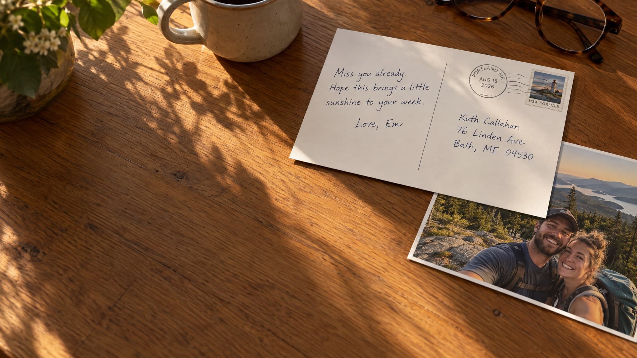 Late afternoon light across a wooden kitchen table: a handwritten postcard addressed to Bath, Maine rests beside a coffee mug and reading glasses, with a printed photo of a couple hiking tucked under its corner.