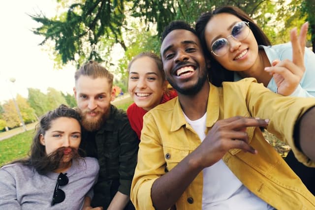 Five friends squeezed into a selfie in a park, one wearing a friend’s hair as a moustache.